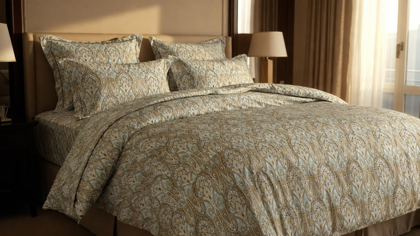 A bed dressed in a beige and light blue patterned comforter set with matching pillows, styled in a warm-toned bedroom with bedside lamps and curtains.