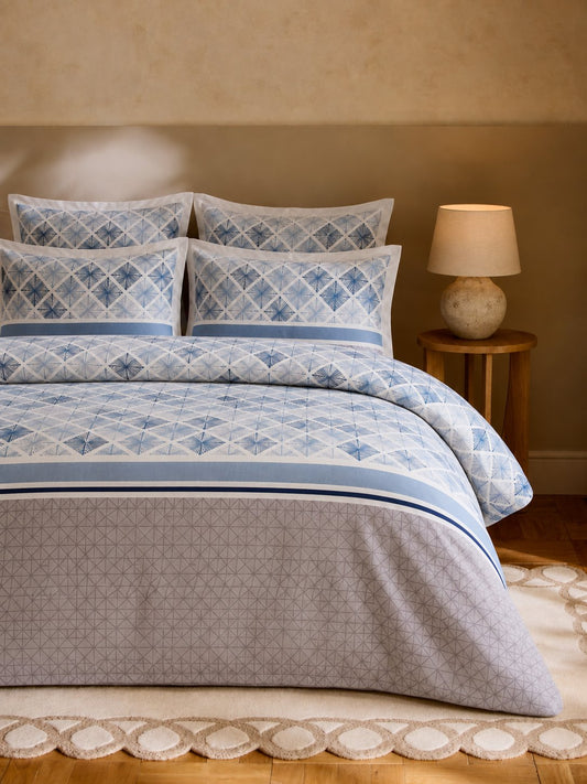 A neatly made bed in a warm-toned bedroom featuring a blue and white geometric patterned bedding set with matching pillows, a light gray patterned comforter, and a wooden nightstand with a lit lamp beside it.