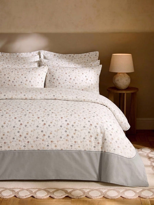 A neatly made bed with a white duvet cover and matching pillow shams featuring a delicate small floral print, finished with a solid light gray bed skirt. The bed is styled in a warm bedroom setting with a wooden nightstand, a lit table lamp, and a patterned rug.
