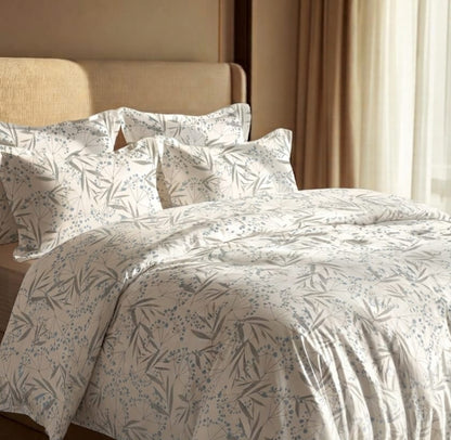 A neatly made bed with a duvet cover and matching pillow shams featuring a delicate blue-gray botanical leaf pattern on a warm off-white background, styled in a bedroom setting with a beige headboard and curtains.