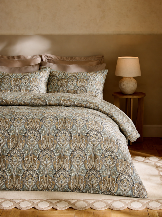 A bed dressed in a patterned duvet cover and matching pillow shams with a paisley design in muted blue, beige, and brown tones, styled in a warm bedroom setting with a bedside lamp.