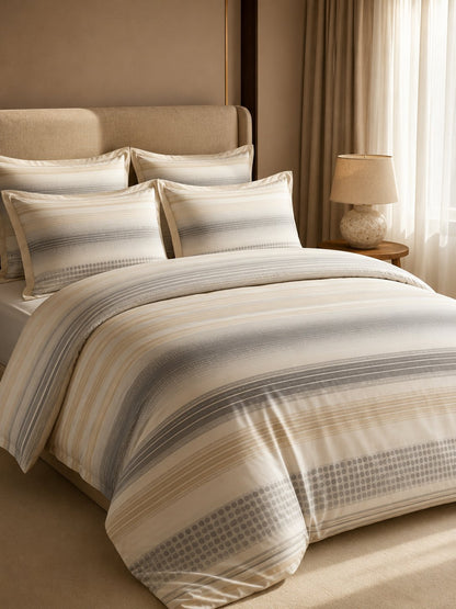 A neatly made bed in a warm-toned bedroom, dressed with a striped duvet cover and matching pillow shams in beige, cream, and gray tones. A bedside lamp sits on a wooden nightstand beside sheer curtains and a beige upholstered headboard.