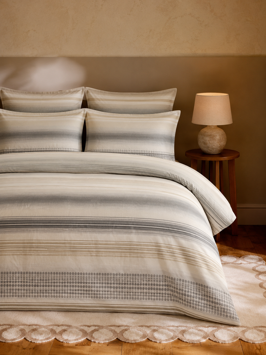 A neatly made bed in a warm bedroom setting, featuring a striped duvet cover and matching pillow shams in beige, gray, and white tones. A wooden nightstand with a lit table lamp sits beside the bed.