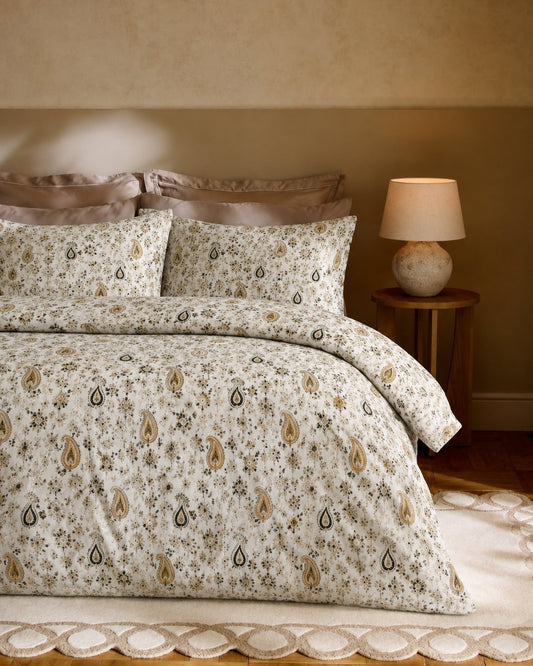A bed dressed in a cream-colored duvet cover and matching pillowcases featuring a delicate floral and paisley pattern in beige, brown, and muted green, styled in a warm, rustic bedroom setting with a wooden nightstand and lamp.