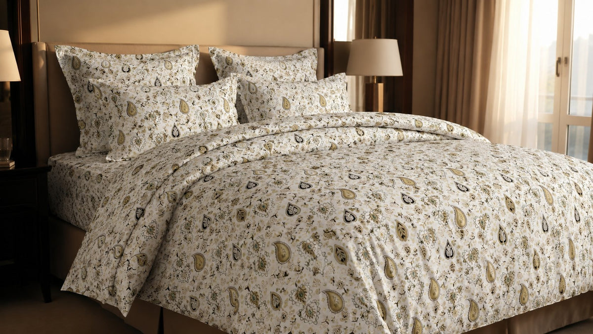 A bed dressed in a cream-colored bedding set with a repeating brown and beige paisley pattern, styled in a warm-toned bedroom setting with bedside lamps and curtains.