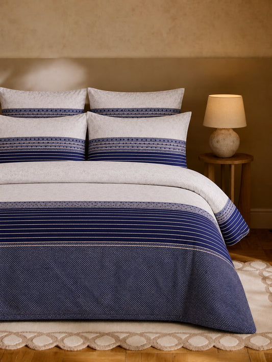 A neatly made bed in a warm bedroom setting, featuring a navy blue and white striped bedding set with matching pillows. A wooden nightstand with a lit lamp sits to the right, and a textured wall and patterned rug are visible in the background.