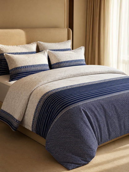 A neatly made bed in a warm beige bedroom setting, featuring a duvet cover and matching pillow shams in cream and navy blue with striped and textured patterns.