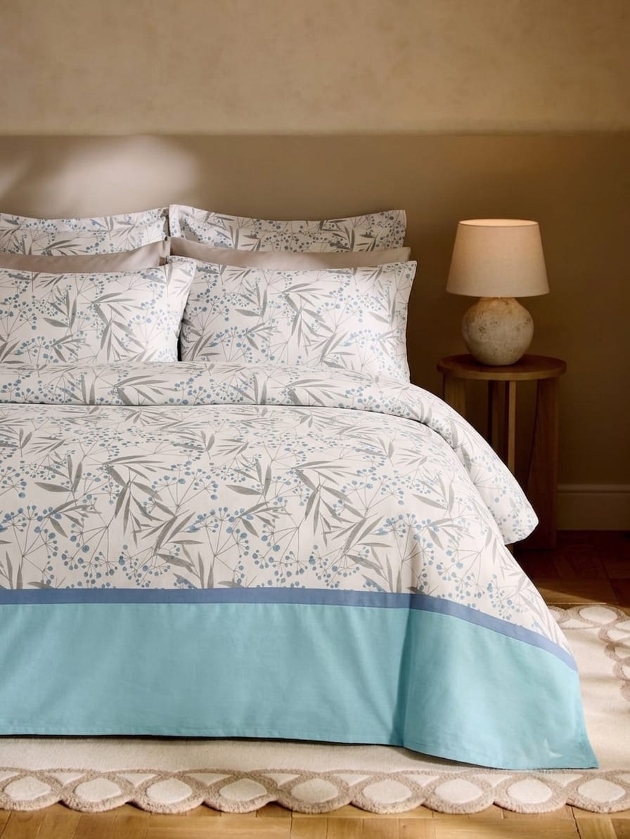 A bed dressed in a white and light blue botanical print bedding set with a solid light blue band at the bottom, styled in a warm bedroom setting next to a wooden nightstand and lamp.