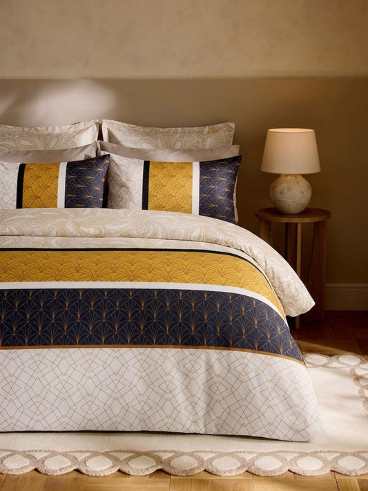 A styled bed dressed in a cream and navy bedding set with geometric and fan-like patterns, including matching pillows, a bedside lamp, and a warm beige wall.