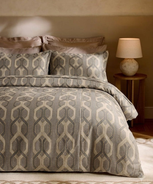 A bed dressed in a patterned beige and gray duvet cover and matching pillow shams, styled in a warm bedroom setting with a wooden side table and lamp.