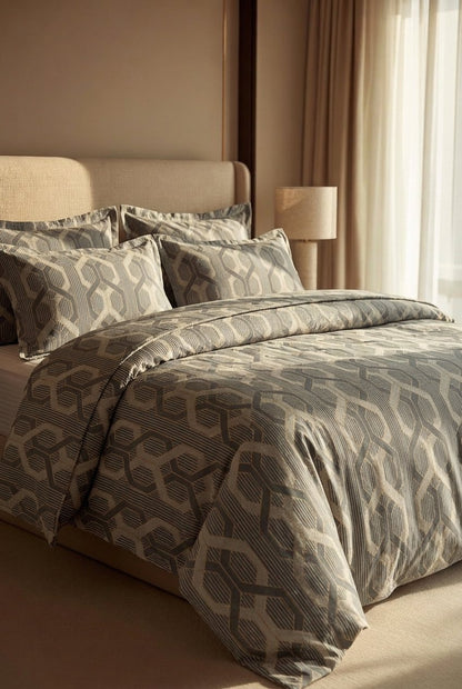 A bed dressed in a gray and beige geometric patterned duvet cover and matching pillow shams, styled in a warm, modern bedroom setting with a beige headboard, curtains, and a bedside lamp.
