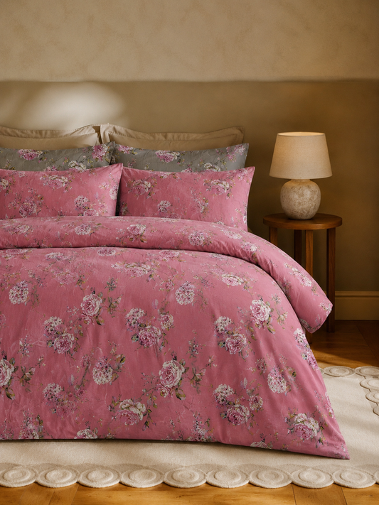 A pink floral duvet cover and pillowcase set styled on a bed in a warm, cozy bedroom setting with a wooden floor, textured wall, and a bedside table with a lamp.