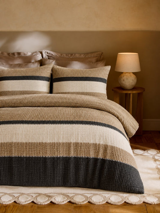 A bed dressed in a textured striped bedding set with beige, cream, and dark charcoal horizontal bands, styled in a warm, softly lit bedroom with a bedside lamp and wood floor.