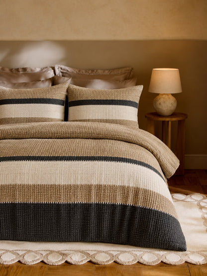 A bed dressed in a textured striped bedding set with beige, cream, and dark charcoal horizontal bands, styled in a warm, softly lit bedroom with a bedside lamp and wood floor.