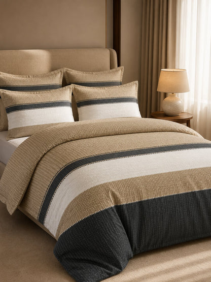 A bed dressed in a striped textured bedding set with a beige headboard, matching pillows, and a duvet featuring horizontal bands of cream, dark charcoal, and tan.