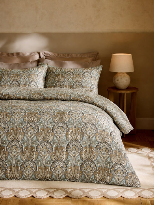 A bed dressed in a patterned duvet cover and matching pillow shams with a paisley design in muted blue, beige, and brown tones, styled in a warm bedroom setting with a bedside lamp.