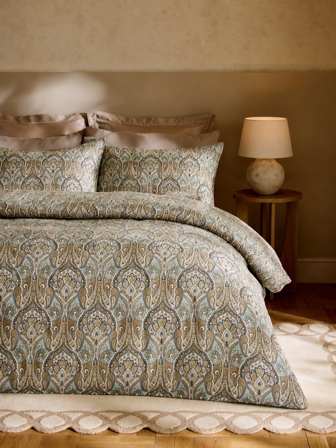 A bed dressed in a patterned duvet cover and matching pillow shams with a paisley design in muted blue, beige, and brown tones, styled in a warm bedroom setting with a bedside lamp.