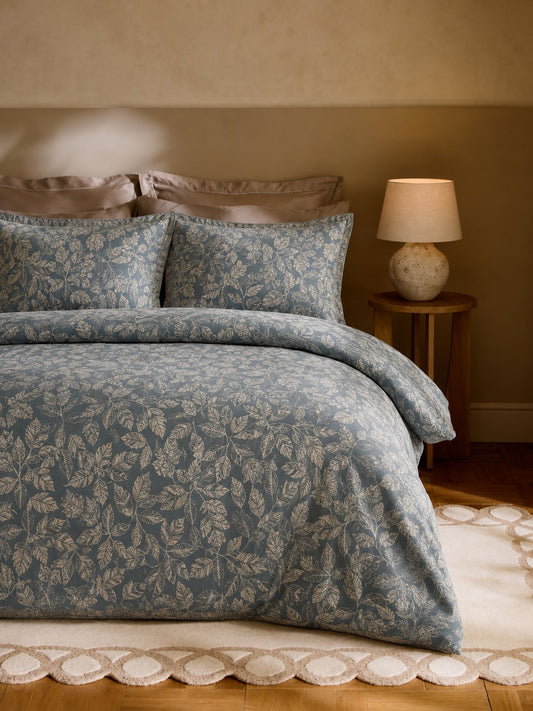 A bed dressed in a blue and beige botanical patterned duvet cover and matching pillow shams, styled in a warm-toned bedroom with a wooden nightstand and lamp.