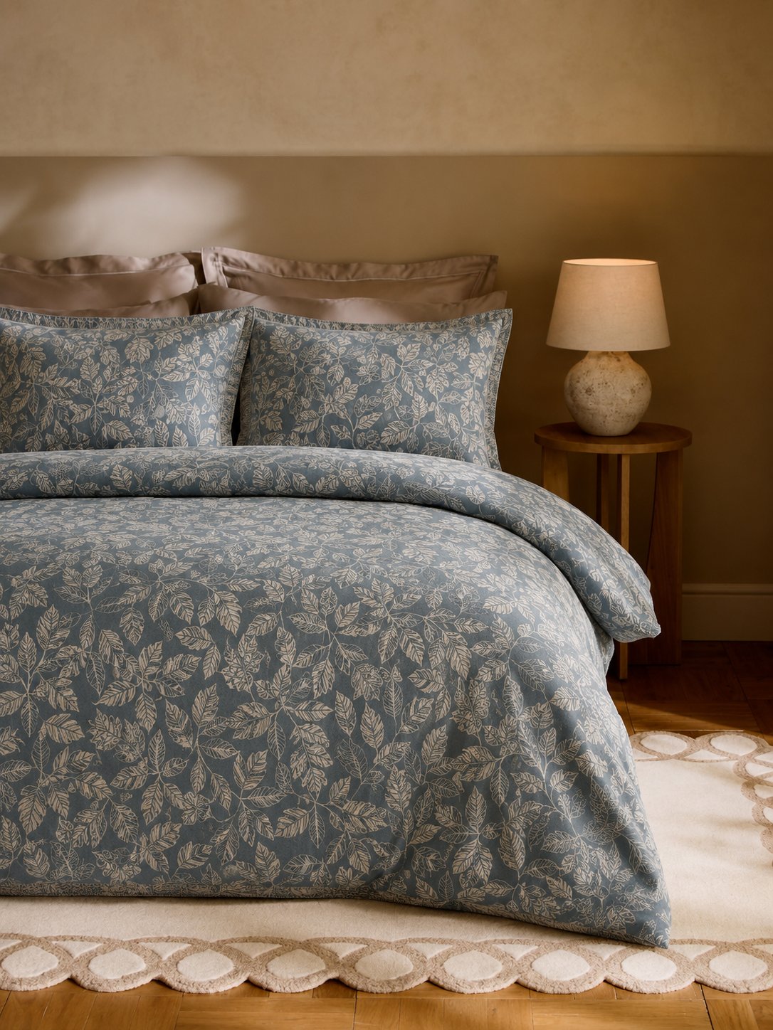A bed dressed in a blue and beige botanical patterned duvet cover and matching pillow shams, styled in a warm-toned bedroom with a wooden nightstand and lamp.
