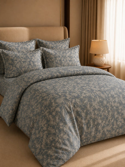 A bed dressed in a muted blue-gray floral-patterned duvet cover and matching pillow shams, styled in a warm beige bedroom with a headboard, curtains, and a bedside lamp.